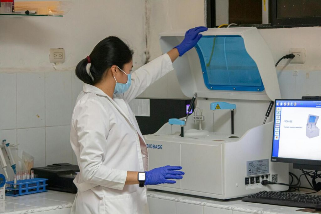 A female laboratory technician wearing a mask, gloves, and lab coat operates a medical analyzer machine next to a computer screen displaying its interface.